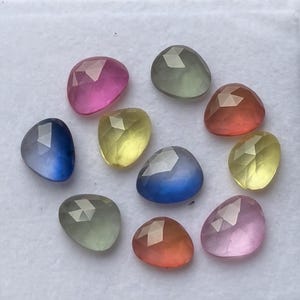 Puede incluir: Una colección de diez piedras preciosas facetadas en forma de pera en varios colores, incluyendo rosa, azul, amarillo, naranja y verde claro. Cada piedra presenta una superficie pulida de múltiples facetas que refleja la luz.