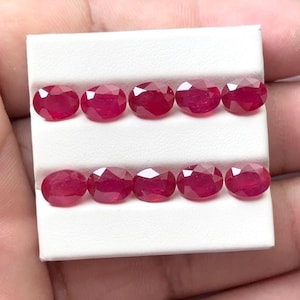 May include: Eight oval-cut, deep red rubies are displayed on a white surface. The gemstones are arranged in two rows of four, showcasing their faceted surfaces and rich color. The rubies are a vibrant, saturated red.