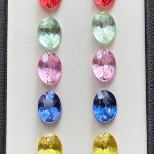 Puede incluir: Una selección de diez gemas de corte ovalado en varios colores, incluyendo rojo, verde, rosa, azul y amarillo. Las gemas se exhiben sobre una superficie blanca, mostrando sus facetas y brillo. Las gemas están dispuestas en dos columnas de cinco.