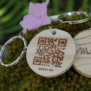 Könnte beinhalten: Ein Holzschlüsselanhänger mit einem silbernen Ring. Auf der einen Seite befindet sich ein QR-Code mit dem Text "misvi.de" und auf der anderen Seite ist der Text "Mi.So." auf einem Holzmaserungshintergrund graviert.