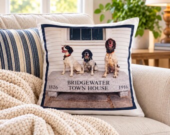 May include: A square throw pillow featuring a photographic print of three dogs sitting on a stone bench in front of a building. The text BRIDGEWATER TOWN HOUSE 1826 1916 is printed on the bench. The pillow has a dark blue border.