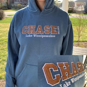 May include: Blue hooded sweatshirt with the embroidered text "CHASE ISLAND Lake Winnipesaukee" in brown and white. The sweatshirt has a front pocket and a drawstring hood. The image shows a close-up of the embroidered text.