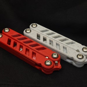 May include: Two butterfly knives, one red and one grey, are displayed against a black background. The knives have a slotted handle design and are held together with silver hardware. The red knife is on the left, and the grey knife is on the right.