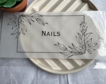 May include: A clear plastic sheet with a black border and floral design. The text "NAILS" is printed in black inside the border.