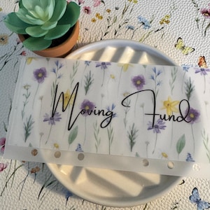 May include: A white, translucent sheet with a floral pattern and the text "Moving Fund" in black script. The sheet has six evenly spaced holes along the bottom edge.