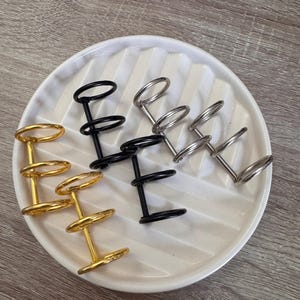 Op de afbeelding: Een verzameling metalen ringhouders in goud, zwart en zilver, geplaatst op een witte, geribbelde keramische schaal. Elke houder heeft een verticale paal met twee of drie ringen. Ontworpen voor naamkaartjes of foto's.