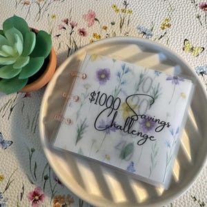 May include: A white and floral patterned journal with a clear plastic cover. The journal has a ring binder and the text "£1000 Savings Challenge" is written in black on the front.