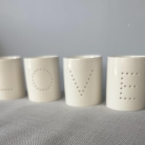 May include: Four cylindrical off-white ceramic candle holders arranged in a row. Each holder features a single letter formed by small, evenly spaced perforations. Together, the holders spell out the word LOVE in a clean, minimalist font style.