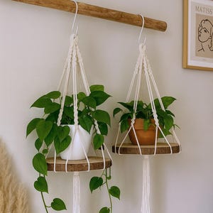 Pode incluir: Duas prateleiras de plantas em macramé com prateleiras de madeira, cada uma com uma planta verde folhosa. Uma planta está num vaso branco, a outra num vaso de terracota. As prateleiras estão suspensas numa viga de madeira.