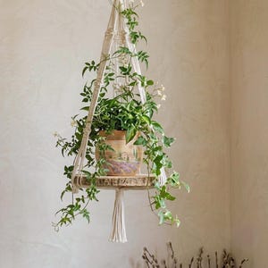 Peut inclure: Une suspension pour plante en macramé faite de corde blanc cassé avec une étagère ronde en bois. Un pot en céramique peint d'un paysage est posé sur l'étagère, contenant une plante verte grimpante à petites fleurs blanches. Un gland pend en bas.