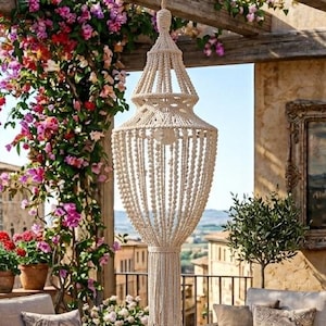 Peut inclure: Scène de patio extérieur avec des meubles en osier, dont un canapé et des fauteuils, disposés autour d'une table basse en bois. Un lustre en macramé est suspendu à une pergola en bois, entouré de plantes grimpantes fleuries et de plantes en pot. Une peinture est accrochée au mur.