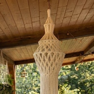 Peut inclure: Une suspension en macramé de couleur crème avec un motif noué complexe et de longues franges, suspendue au plafond en bois d'une véranda au-dessus d'une terrasse en pierre remplie de diverses plantes en pot et fleurs en terre cuite.