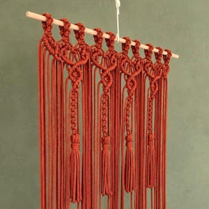 Op de afbeelding: Een roestoranje macramé wandhanger met ingewikkelde knooppatronen, opgehangen aan een lichtgekleurde houten stok. Het ontwerp bevat vier verticale secties met gevlochten details die eindigen in lange, dikke kwastjes.