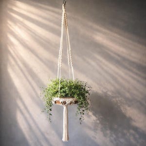 Op de afbeelding: Een macramé plantenbak met een ronde houten basis, met een weelderige groene plant. Het off-white touwontwerp hangt de plantenbak op, waardoor een bohemien esthetiek ontstaat. De hangende bladeren voegen een natuurlijk tintje toe aan de decoratie.