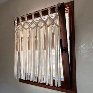 Puede incluir: Cortina de macramé color crema. La sección superior presenta un patrón de diamantes, con largos hilos verticales colgando. Las borlas están espaciadas uniformemente a lo largo del borde inferior. El marco de la ventana es marrón oscuro.
