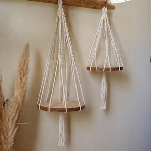 Op de afbeelding: Twee macramé hangende planken met ronde houten platforms en crèmekleurig touw. De planken zijn opgehangen aan een houten balk en tonen een bohemien of rustieke esthetiek. Elke plank heeft een kwast aan de onderkant.