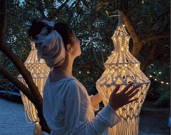 Puede incluir: Dos linternas de macramé en color blanco roto con intrincados patrones de nudos y largos flecos en la base. Cada linterna está iluminada desde el interior por luces cálidas y cuelga de una rama de árbol en un entorno exterior al atardecer.