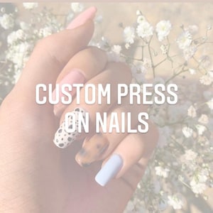May include: Close-up of a hand wearing custom press-on nails with different designs, including a light blue nail, a brown and white marbled nail, and a black and white polka dot nail. The text "CUSTOM PRESS ON NAILS" is visible in the image.