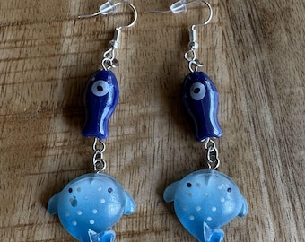 Pode incluir: Um par de brincos pendurados com um pingente em forma de peixe azul escuro com um olho branco, conectado por uma corrente a um pingente de peixe azul claro com bolinhas brancas. Os brincos têm ganchos de orelha prateados.