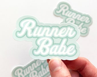 May include: Mint green and white sticker with the words "Runner Babe" in a cursive font. The sticker has a rounded rectangular shape and is held by a hand. Two other stickers are visible in the background.