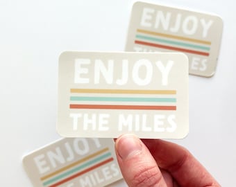 Könnte beinhalten: Beige rechteckige Aufkleber mit abgerundeten Ecken. Der Text "ENJOY THE MILES" ist in Weiß über horizontalen gelben, blauen und roten Streifen angeordnet.