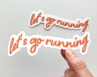 May include: Two white stickers with the text "let's go running" in orange script. The text has a shadow effect, and one sticker is being held by a person's fingers. The stickers are on a white background.