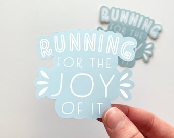 Könnte beinhalten: Zwei hellblaue Aufkleber mit dem weißen Schriftzug "RUNNING FOR THE JOY OF IT". Ein Aufkleber wird von einer Hand gehalten. Die Aufkleber haben eine wolkenartige Form mit weißen Akzenten.