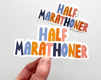 以下が含まれることがあります： 遊び心のあるフォントで「HALF MARATHONER」というテキストが書かれたカラフルなステッカー。ステッカーは白い背景に青、オレンジ、ピンクの文字で構成されています。1枚のステッカーが手で持たれており、もう1枚は背景にあります。