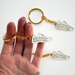 Puede incluir: Tres llaveros con anillos y cadenas doradas. Cada llavero tiene un dije blanco en forma de zapato con el texto "BEST RUNNING BUDDY EVER" en negro. Los llaveros se sostienen en una mano.