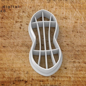 Puede incluir: Cortador de galletas de plástico blanco en forma de cacahuete con un diseño detallado. El cortador está sobre una superficie de madera marrón.