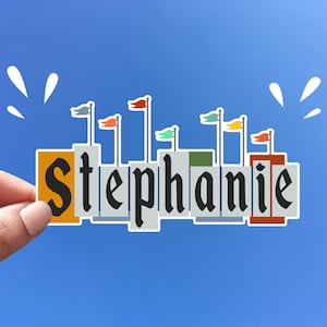 Pode incluir: Um adesivo com o nome "Stephanie" escrito em letras pretas sobre um fundo branco. Cada letra &eacute; cercada por uma borda cinza e tem uma pequena bandeira no topo. As bandeiras s&atilde;o vermelhas, laranja, amarelas, verdes, azuis e roxas.