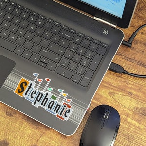 Pode incluir: Um computador port&aacute;til preto com um adesivo que diz "Stephanie" na tampa. O laptop est&aacute; em uma superf&iacute;cie de madeira com um mouse sem fio preto ao lado.