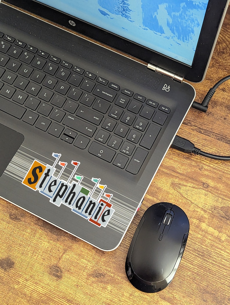 Pode incluir: Um computador port&aacute;til preto com um adesivo que diz "Stephanie" na tampa. O laptop est&aacute; em uma superf&iacute;cie de madeira com um mouse sem fio preto ao lado.