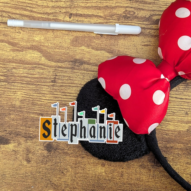 Pode incluir: Uma faixa de cabelo com um la&ccedil;o vermelho de bolinhas brancas e uma base de feltro preto. A faixa de cabelo tem uma etiqueta com o nome "Stephanie" e um gr&aacute;fico de castelo.