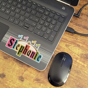 Pode incluir: Um laptop preto com um adesivo colorido que diz "Stephanie" na tampa. O laptop est&aacute; em uma superf&iacute;cie de madeira com um mouse sem fio preto ao lado.