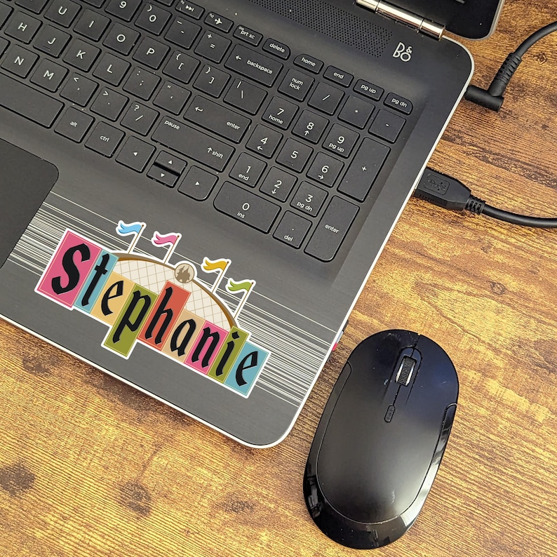 Pode incluir: Um laptop preto com um adesivo colorido que diz "Stephanie" na tampa. O laptop est&aacute; em uma superf&iacute;cie de madeira com um mouse sem fio preto ao lado.