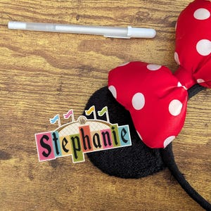 Pode incluir: Uma faixa de cabe&ccedil;a Minnie Mouse de bolinhas vermelhas e brancas com uma orelha preta e um adesivo colorido que diz "Stephanie".
