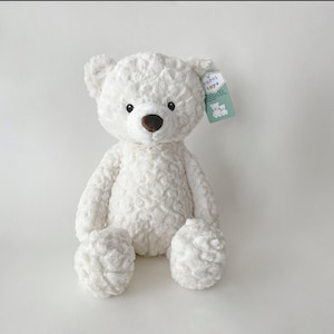 May include: White plush teddy bear with a brown nose and black eyes. The bear is sitting with its legs crossed and has a tag that says "curci toys" on its right ear.