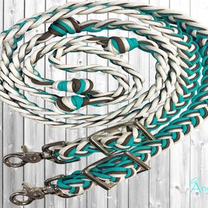 Peut inclure: Rênes de cheval tressées en turquoise, blanc et marron. Les rênes sont dotées d'une quincaillerie argentée, comprenant deux boucles et deux mousquetons. Le design incorpore un motif répétitif des trois couleurs, créant un accessoire visuellement attrayant.