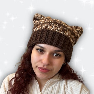 May include: A brown and beige crocheted beanie with cat ears. The beanie has a brown brim and a beige and brown patterned top.