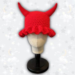 May include: A bright red, crocheted hat with two curved horns and a ruffled brim. The hat is displayed on a mannequin head against a soft, star-speckled background. This unique hat is a fun accessory.