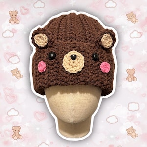 May include: Brown crocheted bear hat with pink cheeks and black eyes. The hat is on a brown mannequin head.