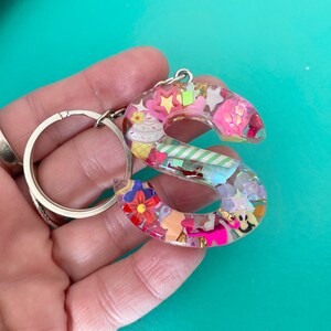 May include: A pink and white resin keychain shaped like the letter S. The letter is filled with colorful confetti, sprinkles, and other small decorations.