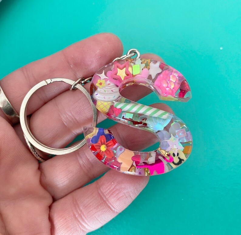 May include: A pink and white resin keychain shaped like the letter S. The letter is filled with colorful confetti, sprinkles, and other small decorations.