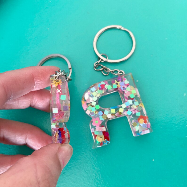 May include: Two resin keychains with glitter and colorful confetti. One keychain is the letter R and the other is a circle.