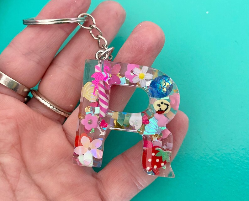 May include: A pink, blue, and yellow resin letter R keychain with a silver key ring. The letter is filled with colorful glitter, beads, and charms.