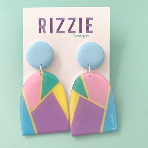 May include: A pair of colorful geometric earrings with a light blue circle at the top of each earring. The earrings are made of resin and have a glossy finish. The earrings are shaped like an arch and have a mix of pink, yellow, purple, blue, and green colors.