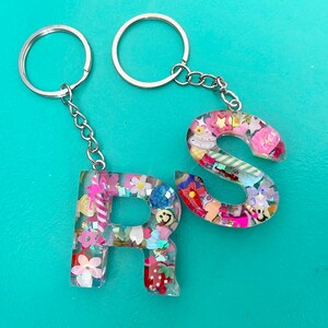 May include: A silver keychain with two key rings and two resin letter charms. The charms are shaped like the letters "R" and "S" and are filled with colorful confetti and glitter.