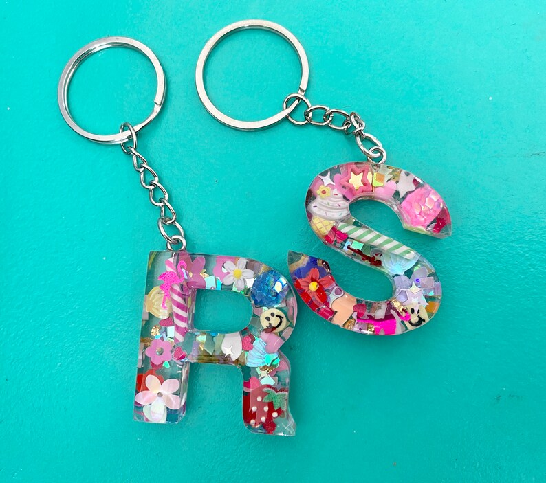 May include: A silver keychain with two key rings and two resin letter charms. The charms are shaped like the letters "R" and "S" and are filled with colorful confetti and glitter.