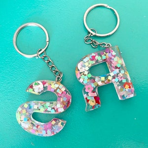 May include: Two keychains with silver key rings and chains. The charms are clear resin letters filled with colorful glitter and confetti. The letters are "S" and "R".